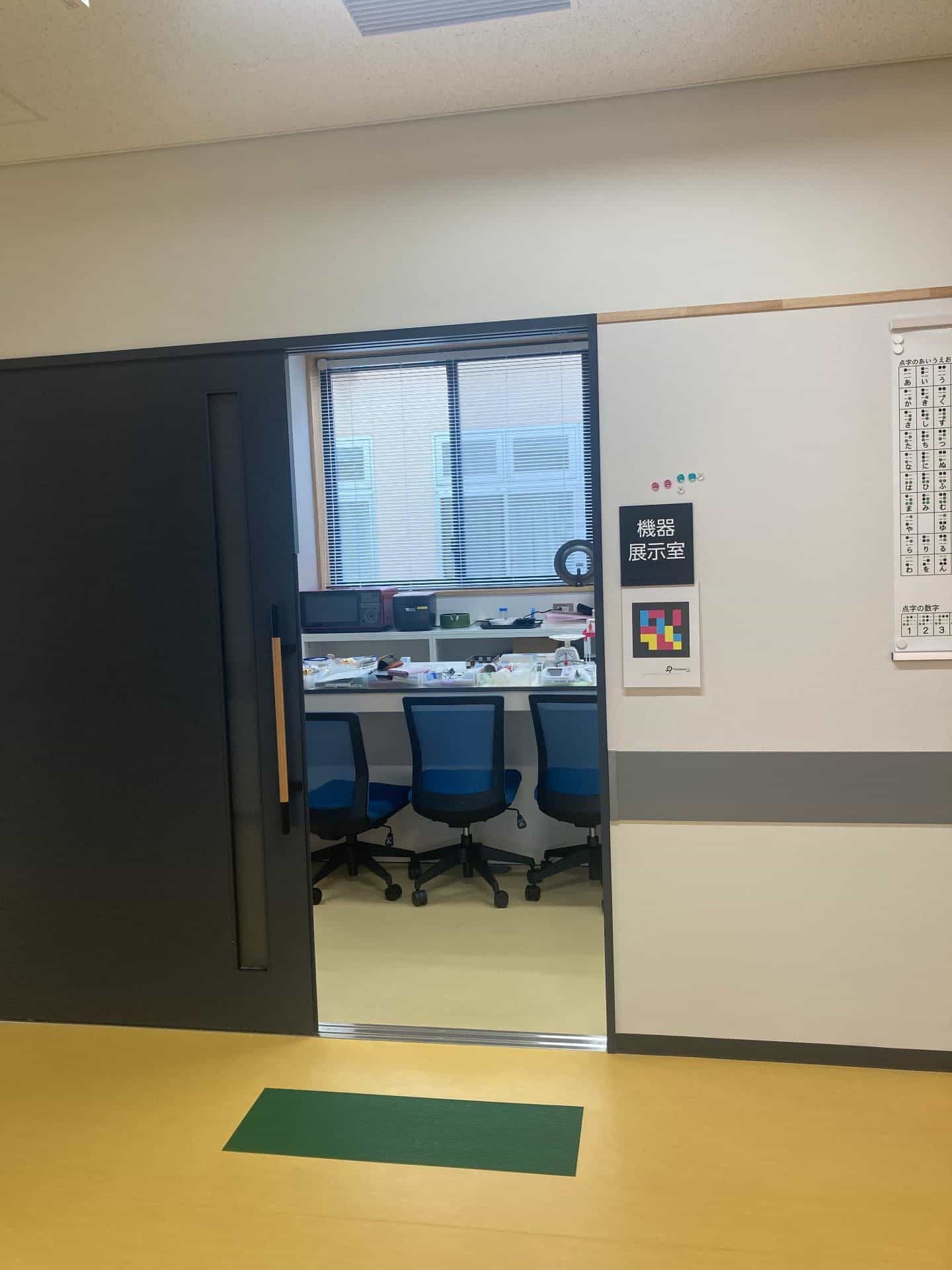 Porta scorrevole scura socchiusa della sala espositiva degli ausili («機器展示室»): all’interno si vede un lungo tavolo bianco continuo con sedie da ufficio blu e, sul tavolo, dispositivi di supporto per persone con disabilità visiva —stampante braille, videoingranditore con anello luminoso, microonde, prodotti di tiflotecnologia—; sulla parete destra, targa tattile nera con «機器展示室» e, sotto, il cartello A4 del codice NaviLens GO; alla parete, un poster appeso con la tavola del braille «点字のあいうえお»; pavimento giallo con tappeto verde