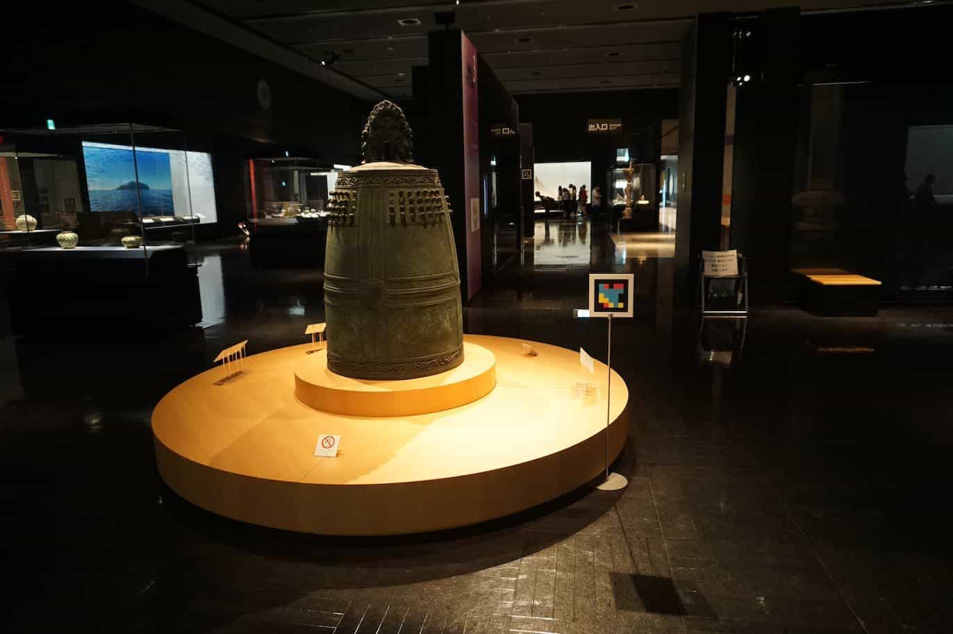 九州国立博物館で、照明の当たる台の上の大きな青銅の鐘。キャプションの横にNaviLensコード
