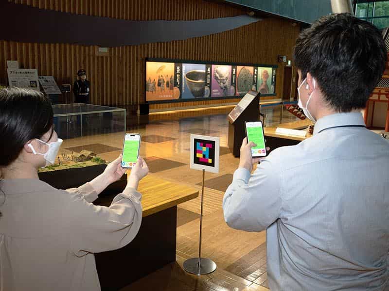 九州国立博物館の文化交流展示室で、2人の来館者がスタンドに設置されたNaviLensコードにスマートフォンをかざしている様子。奥には大きな展示パネル