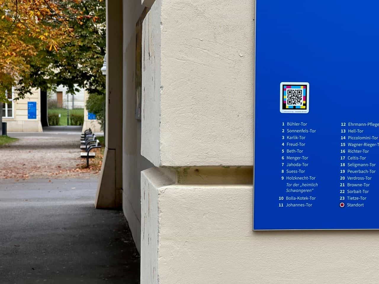 Schräge Seitenansicht der blauen Tafel der Tore der Erinnerung mit vollständiger Liste der 23 Tore in zwei Spalten und darüber dem in Weiß gerahmten NaviLens-Code