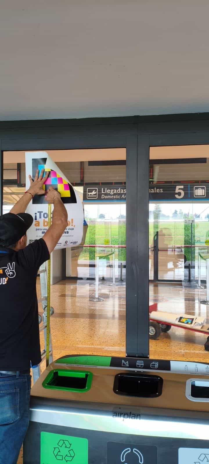 Uniformierter Airplan-Mitarbeiter auf einer Leiter befestigt einen quadratischen NaviLens-Code in Cyan, Magenta, Gelb und Rot auf einem weißen Schild «¡Todos a bordo!, für ein inklusiveres Reiseerlebnis»; im Hintergrund die verglasten automatischen Türen des Zugangs «Llegadas Nacionales 5 / Domestic Arrivals» am Flughafen José María Córdova mit drei Airplan-Recyclingbehältern im Vordergrund