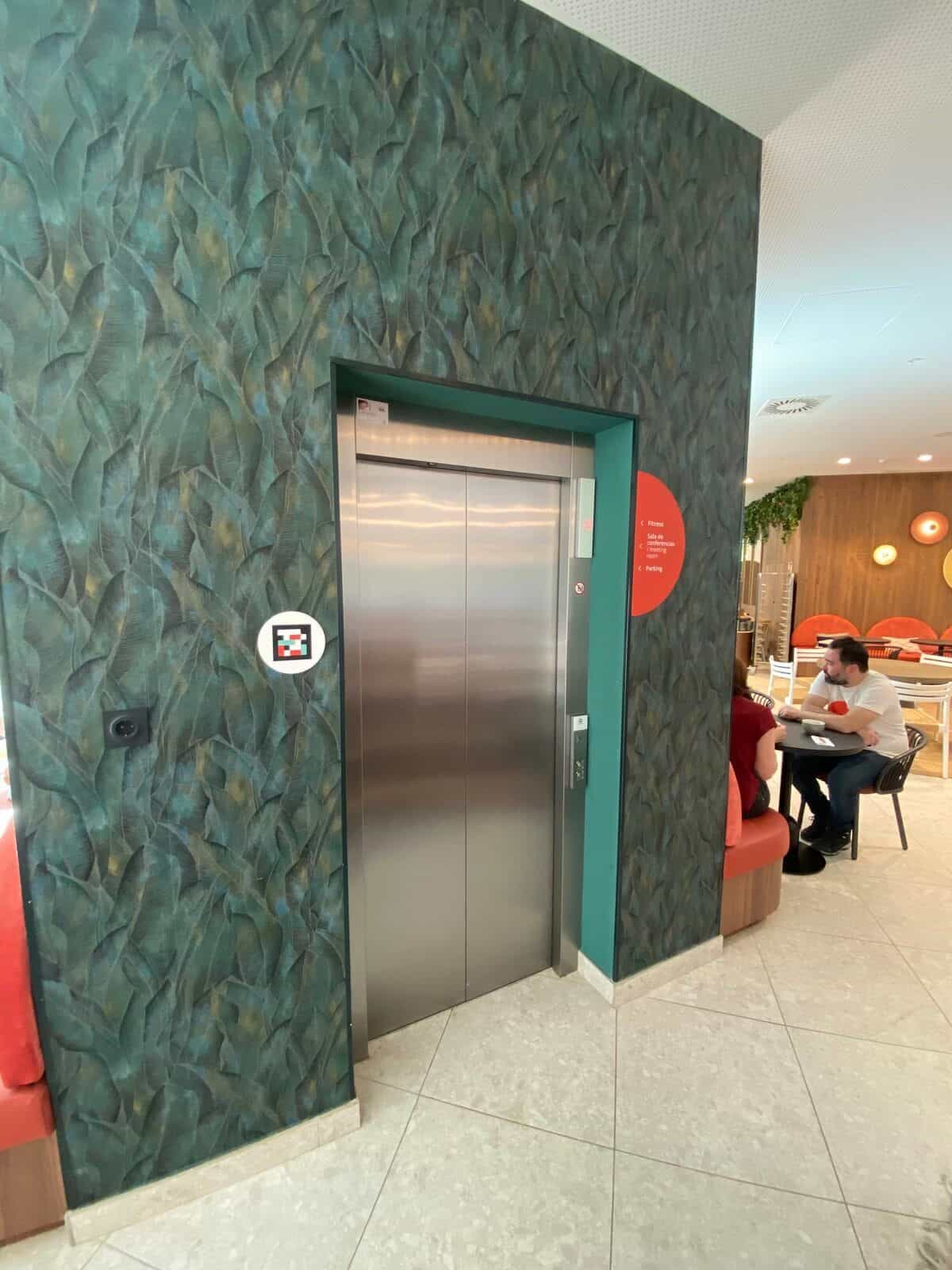 Erdgeschoss-Aufzug am ibis Styles Sevilla in eine Wand mit tropisch-grüner Tapete eingelassen: oranges Rundschild „Fitness · Konferenzraum · Parkhaus“ und weißer Disc mit NaviLens-Code links