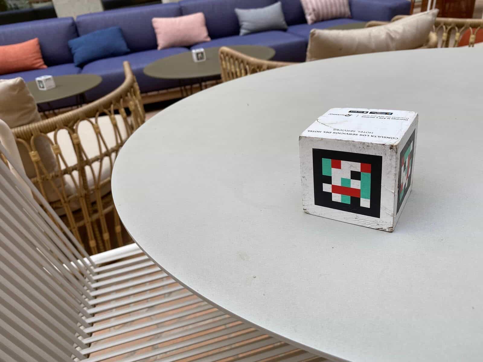 Runder weißer Loungetisch am ibis Styles Sevilla mit NaviLens-Würfel darauf; im Hintergrund großes blaues Sofa mit bunten Kissen