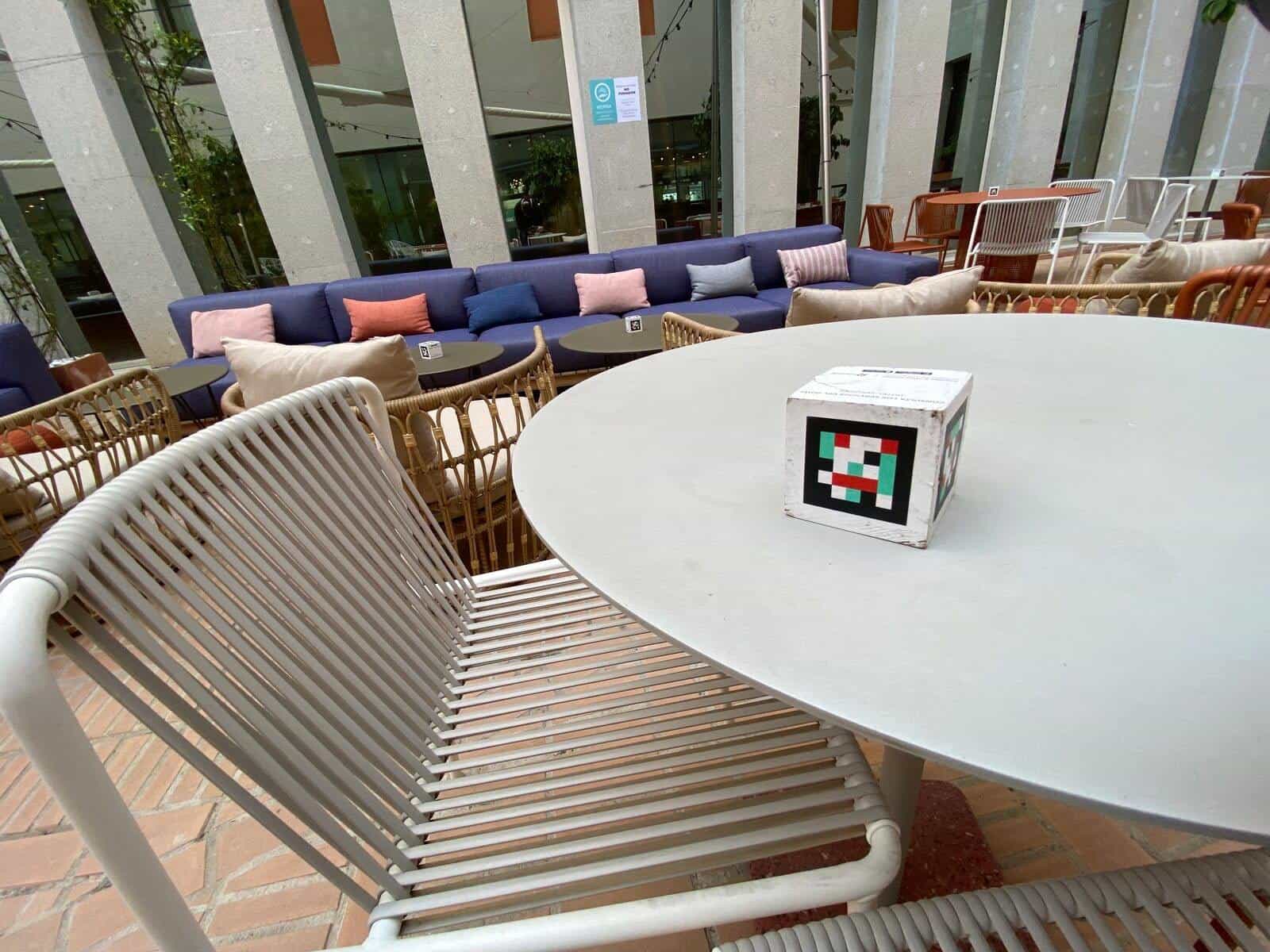 Wide view of the lounge-bar at ibis Styles Sevilla with tall concrete columns, a large blue sofa with colourful cushions and round tables, each with a NaviLens cube