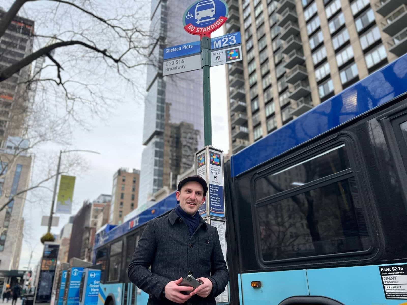 Viaggiatore con il telefono accanto al palo della fermata M23-SBS «Chelsea Piers · E 23 St & Broadway» a Manhattan, con vari codici NaviLens GO e i cartelli «For bus arrival and service info — Scan with NaviLens GO», «Pay at fare machines before boarding» e «Paper ticket required for Select Bus Service»; a fianco, un bus azzurro +SelectBusService