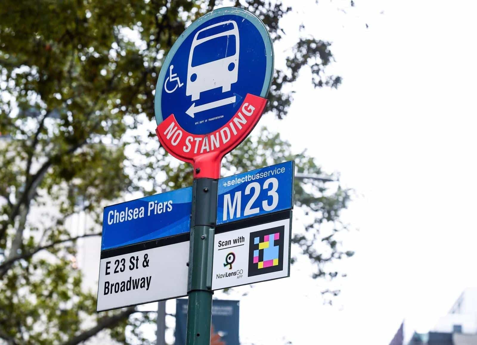 Close-up of the “No Standing” pole at the M23-SBS “Chelsea Piers · E 23 St & Broadway” stop: below the blue line sign, an official white “Scan with NaviLens GO app” plate with the colourful code and the arrow showing the direction of the M23 Select Bus Service