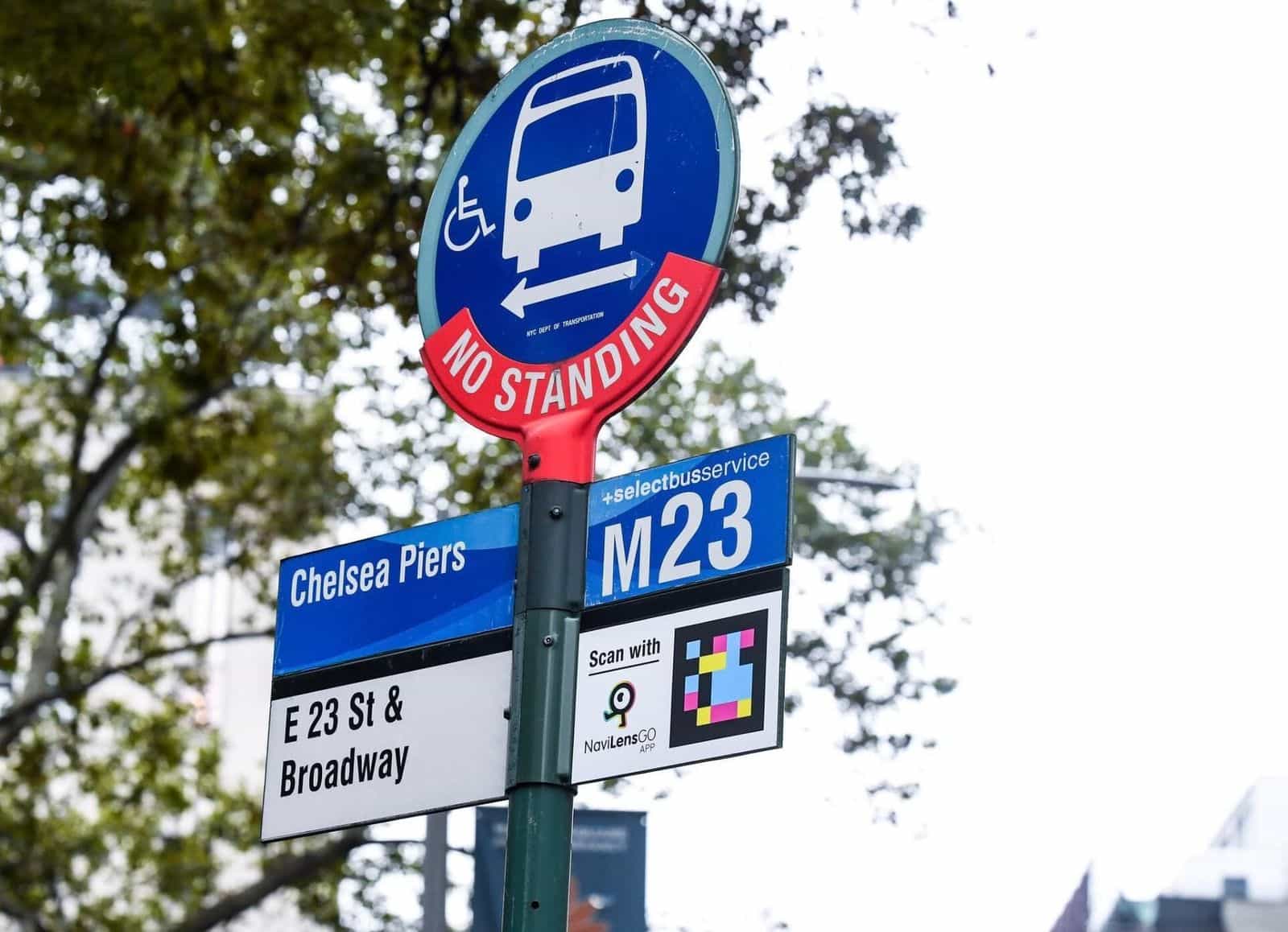 Dettaglio del palo «No Standing» della fermata M23-SBS «Chelsea Piers · E 23 St & Broadway»: sotto il cartello azzurro della linea, una targa bianca ufficiale «Scan with NaviLens GO app» con il codice a colori e la freccia che indica il senso di marcia del bus M23 Select Bus Service