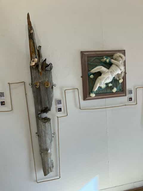 Parete bianca con due opere: a sinistra, un pezzo verticale di legno di mare in forma di maschera allungata con due occhi gialli inseriti; a destra, la scultura tessile della figura bianca incorniciata in legno scuro; fra le due, la corda di canapa percorre la parete a zig-zag