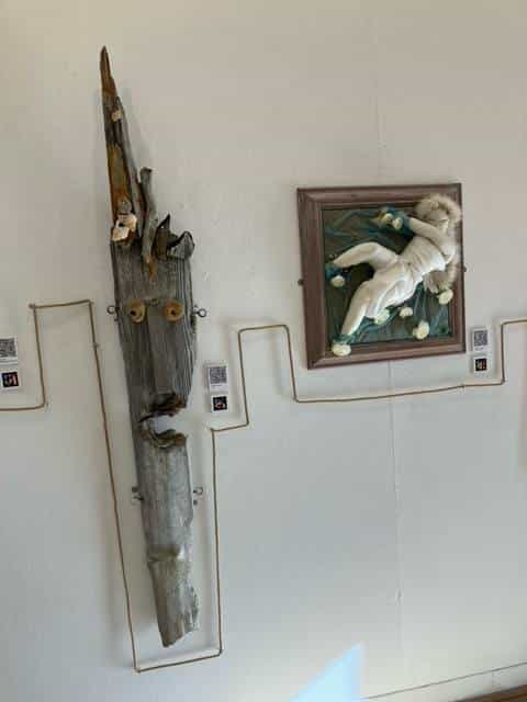 White wall with two works: on the left, a vertical drift-wood piece shaped as an elongated mask with two yellow nailed eyes; on the right, the textile figure framed in dark wood; between them, the hemp rope zig-zags across the wall