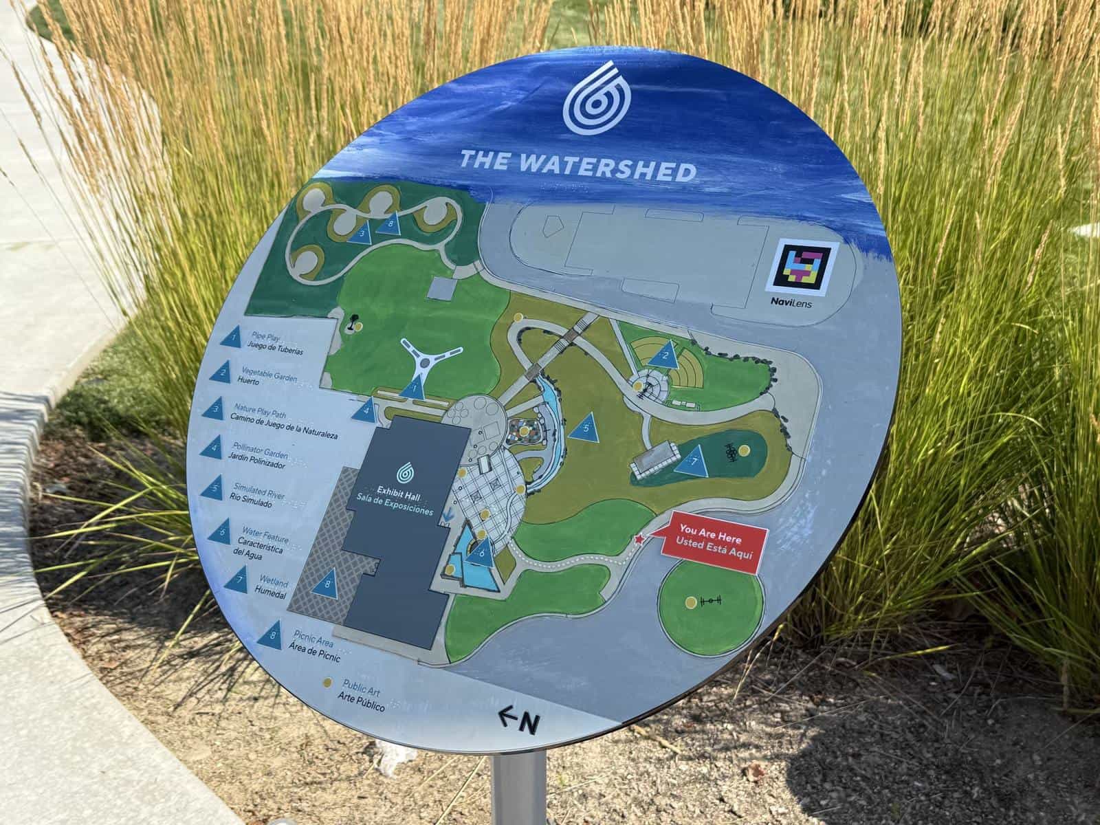 The WaterShed's round site map on a post among ornamental grasses: the “THE WATERSHED” wordmark, the campus plan with its bilingual legend — Pipe Play / Juego de Tuberías, Exhibit Hall / Sala de Exposiciones, Wetland / Humedal — and, top right, the NaviLens code with its label