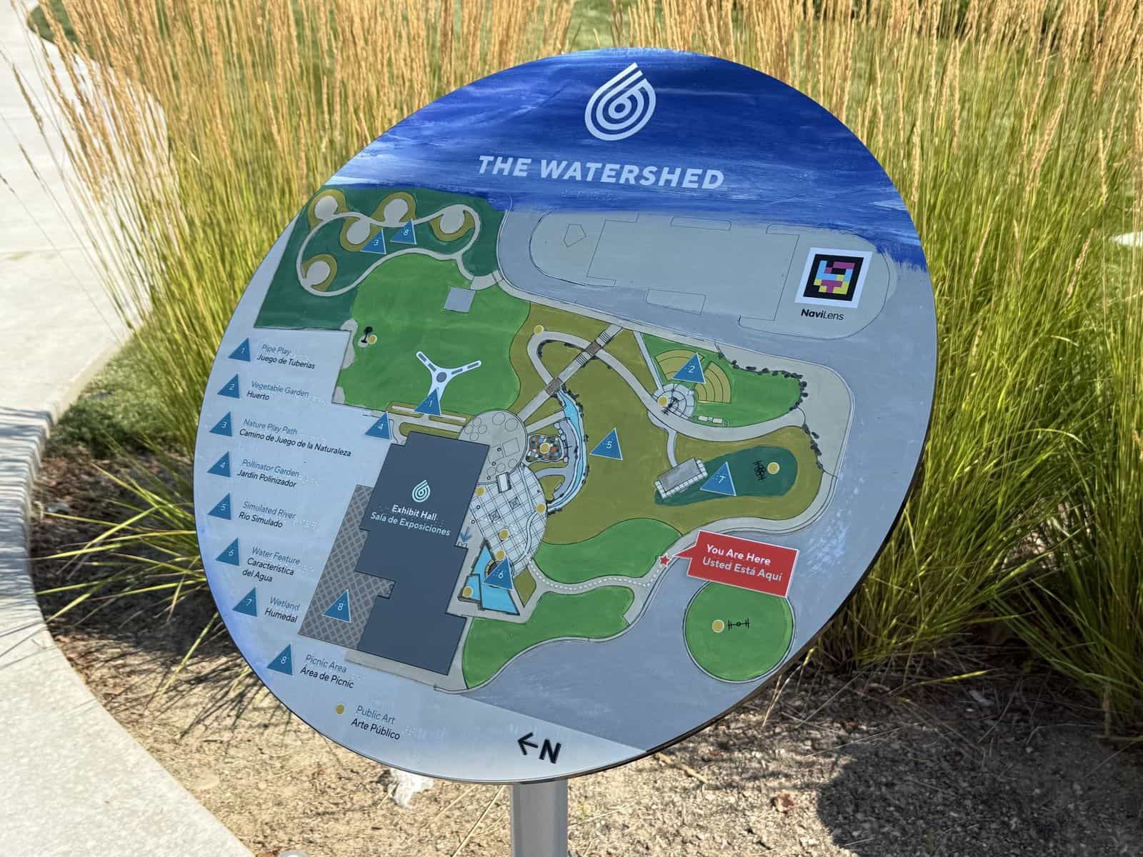 Runde Übersichtskarte von The WaterShed auf einem Pfosten zwischen Ziergräsern: der Schriftzug „THE WATERSHED“, der Geländeplan mit zweisprachiger Legende – Pipe Play / Juego de Tuberías, Exhibit Hall / Sala de Exposiciones, Wetland / Humedal – und oben rechts der NaviLens-Code mit seiner Beschriftung