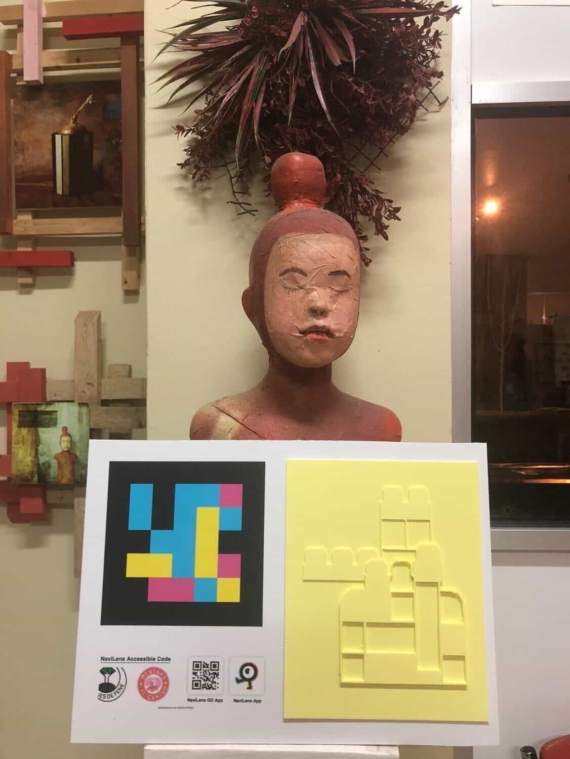 Panel del proyecto «Tocando as Meninas»: a la izquierda código NaviLens cuadrado en cian, magenta y amarillo con sellos de IES de Fene, Meninas de Canido y NaviLens GO; a la derecha relieve amarillo en 3D de una Menina con figuras geométricas. Detrás, busto de cerámica del estudio del artista