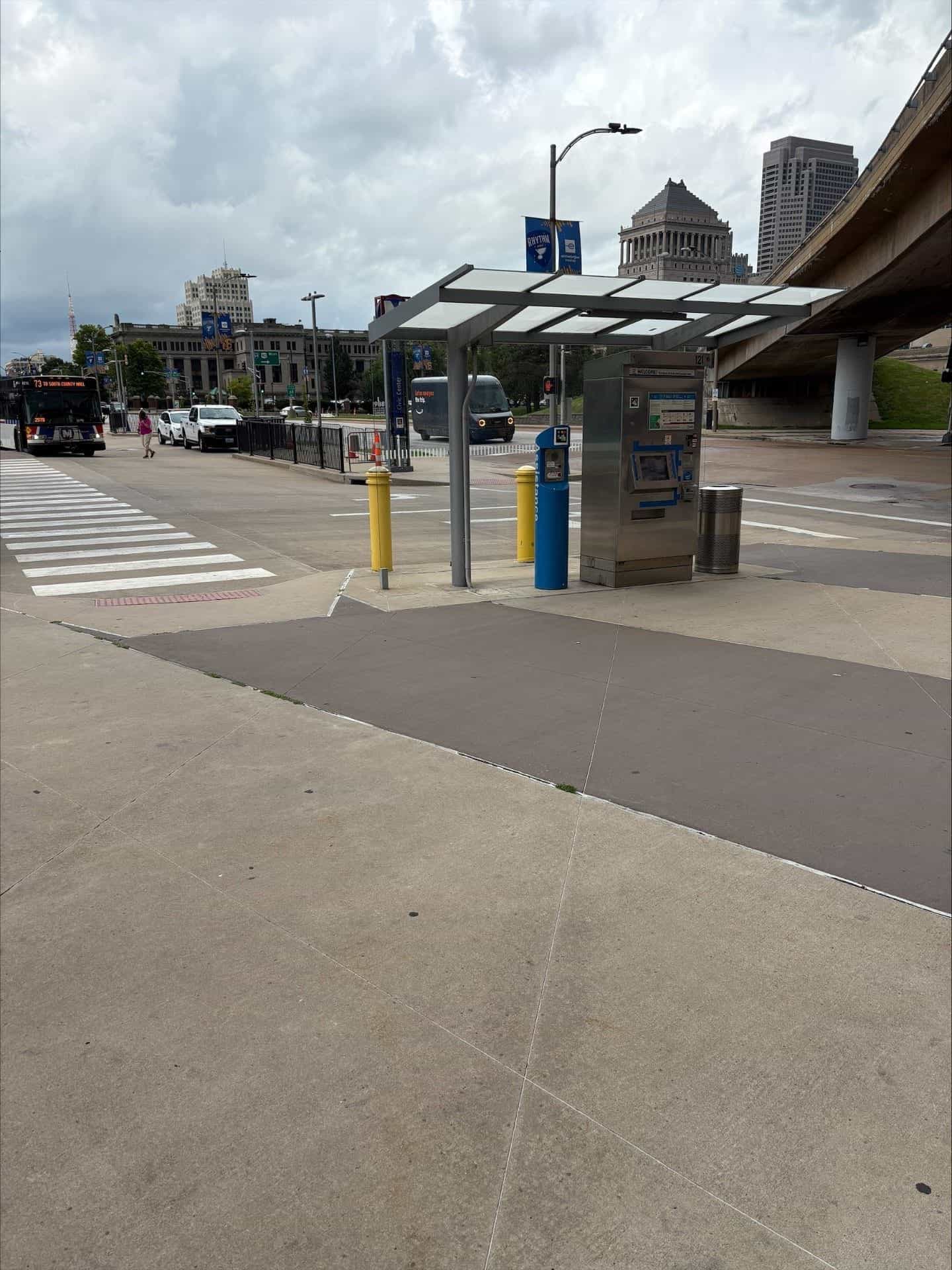 Close-up of the stainless steel ticket vending machine (TVM) under the white canopy: a NaviLens code in yellow, pink, blue and black squares is fixed to the front, next to the blue "Assistance" help post; in the background, the dome of a historic downtown building, a blue "RHYTHM" banner hanging from the lamp post and the concrete overhang of the MetroLink walkway