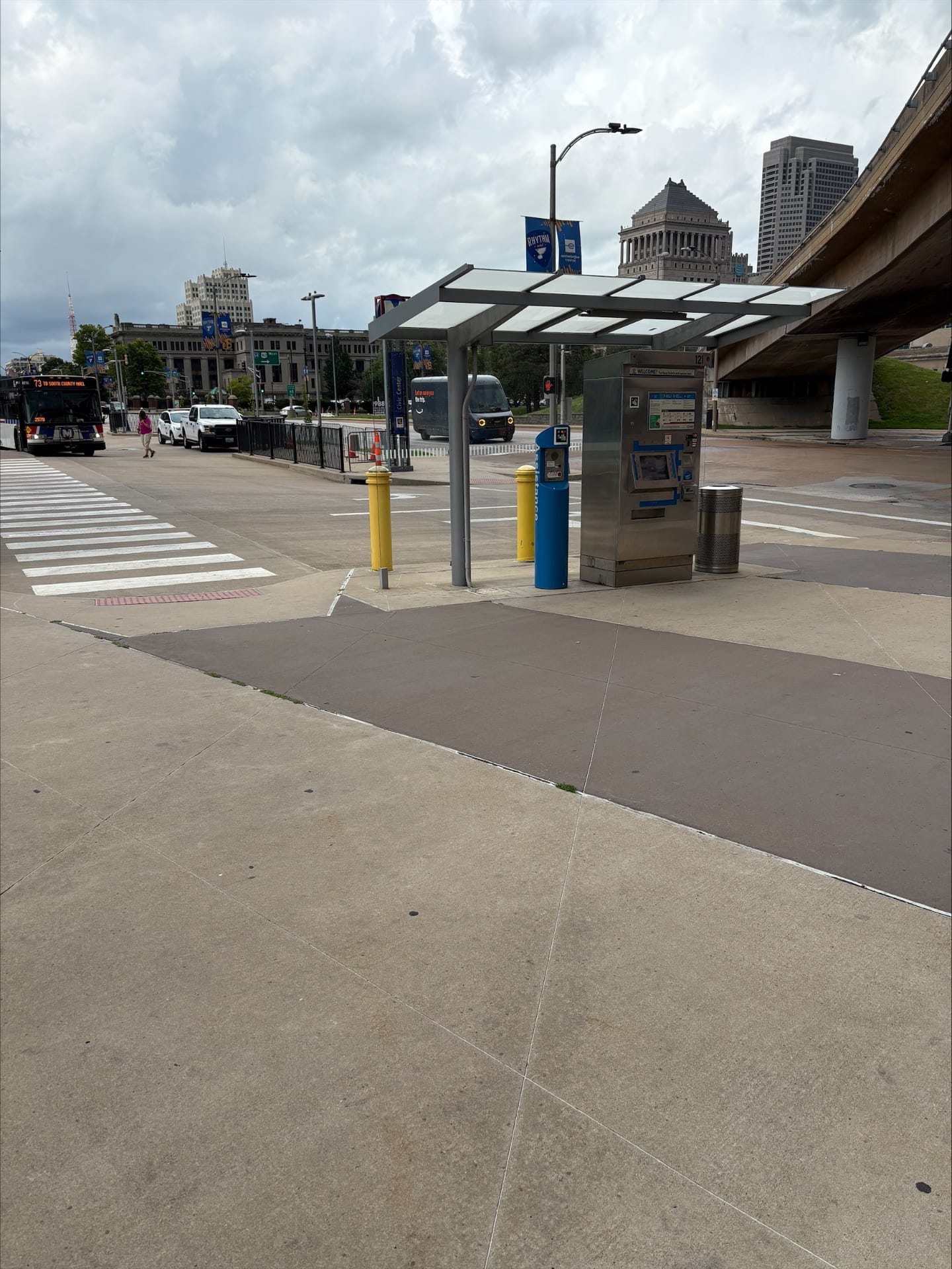 Close-up of the stainless steel ticket vending machine (TVM) under the white canopy: a NaviLens code in yellow, pink, blue and black squares is fixed to the front, next to the blue "Assistance" help post; in the background, the dome of a historic downtown building, a blue "RHYTHM" banner hanging from the lamp post and the concrete overhang of the MetroLink walkway