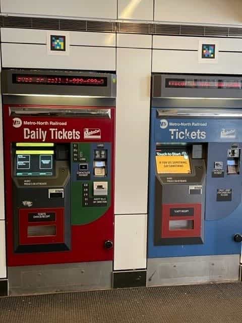 Two Metro-North Railroad ticket machines —«Daily Tickets» in red and «Tickets» in blue— each with a NaviLens code on top, next to the LED info screen