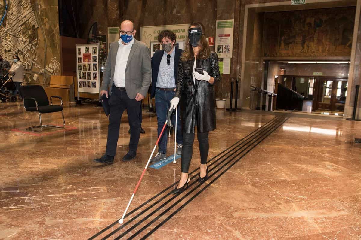 La consejera María Navarro recorre con antifaz y bastón blanco la franja podotáctil instalada por Implaser sobre el suelo de mármol del hall del Ayuntamiento de Zaragoza