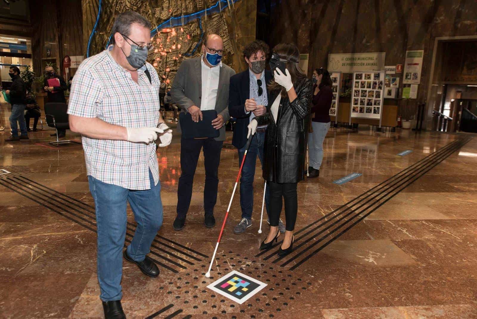 María Navarro, wearing a blindfold and white gloves, raises her phone to read a colourful square NaviLens code stuck on a tactile paving cross on the floor of the Zaragoza City Hall lobby; three companions watch the demonstration