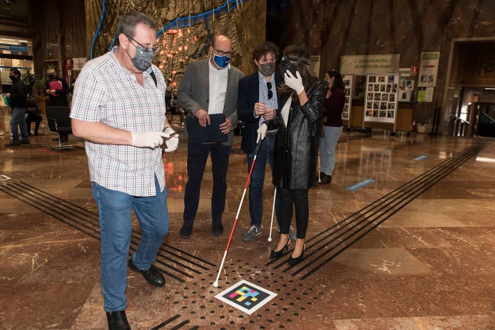 María Navarro, con mascherina oscurante e guanti bianchi, alza il telefono per leggere un codice NaviLens quadrato a colori applicato su una croce tattile sul pavimento della hall del Comune di Saragozza; tre accompagnatori osservano la dimostrazione