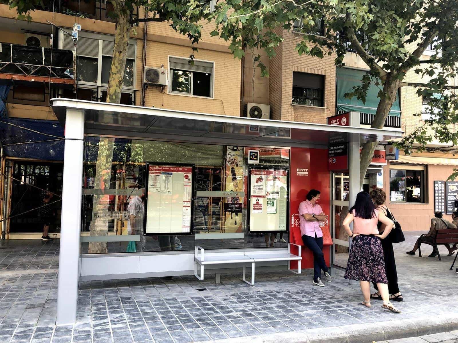 Front view of the EMT València shelter 'Amado Granell Mesado (odd) – la Plata' with zonal map info panels, lines 35 and N9; a traveller leaning on the red side module reads the panel and, at the top of the glass, a colourful square NaviLens code stands out
