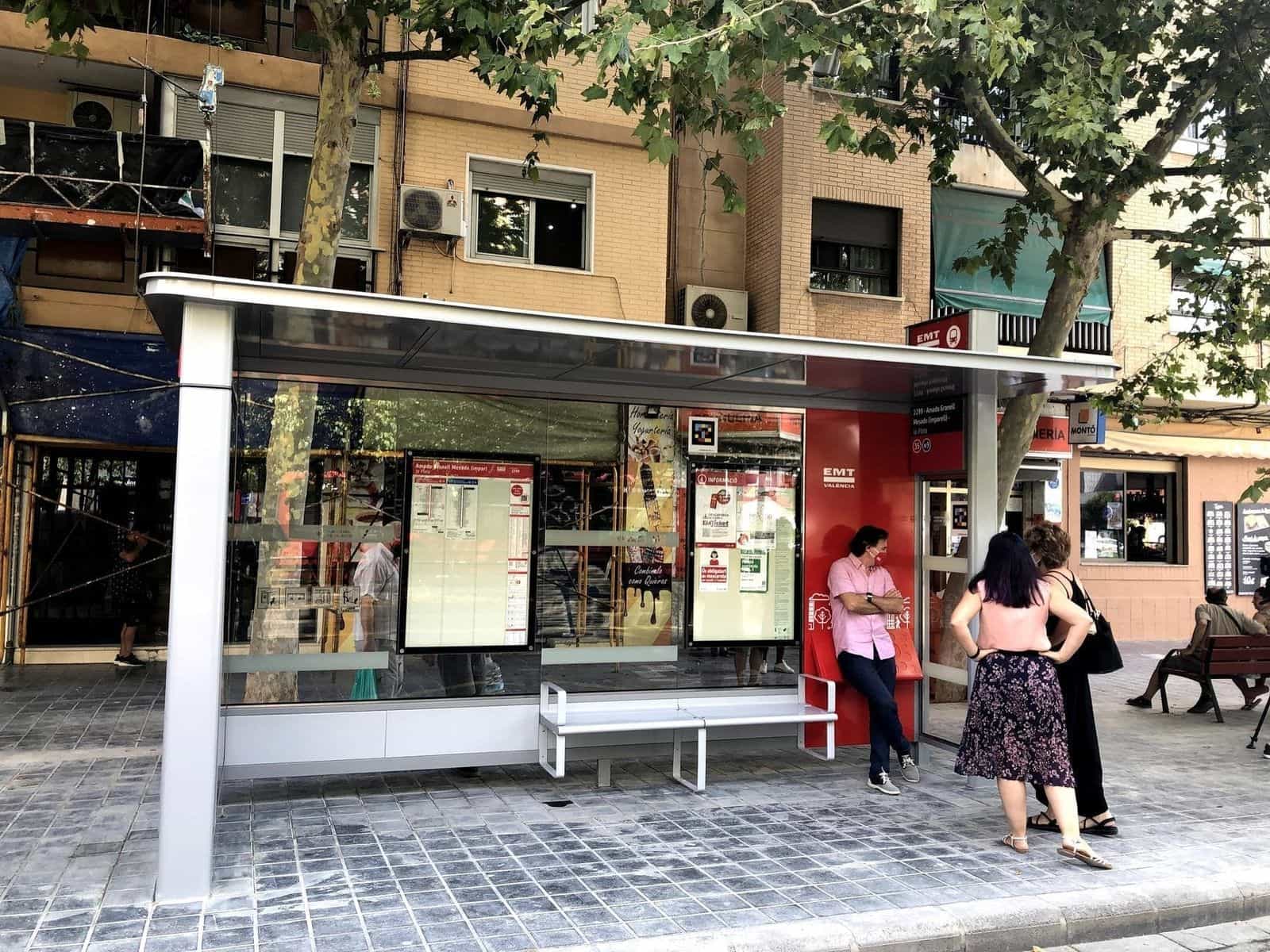 Veduta frontale della pensilina della EMT València «Amado Granell Mesado (impar) – la Plata» con i pannelli informativi del plànol zonal, linee 35 e N9; un viaggiatore appoggiato al modulo rosso laterale consulta il pannello e, nella parte alta del vetro, si distingue un codice NaviLens quadrato a colori