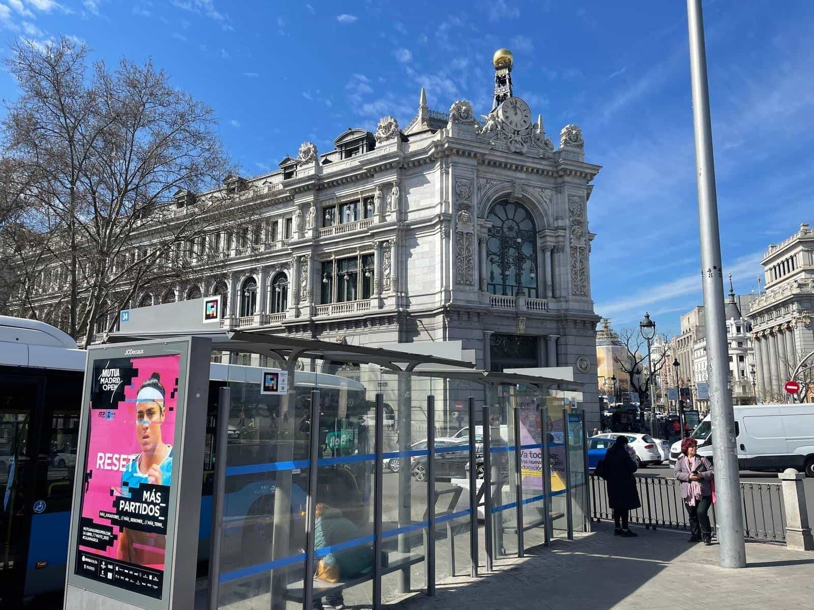 EMT shelter with the numeric plate “34” in front of the Banco de España building on Calle Alcalá; on the roof a colourful NaviLens code stands out next to the stop sign