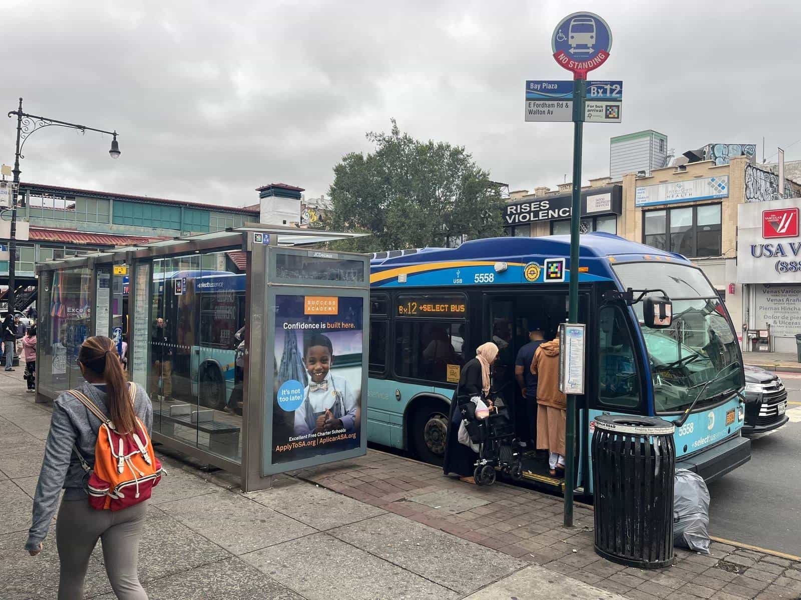 Bx12-SBS shelter at “Bay Plaza · E Fordham Rd & Walton Av” with a NaviLens code next to the “For bus arrival” sign; in the background, coach 5558 with doors open and riders boarding, including a wheelchair user