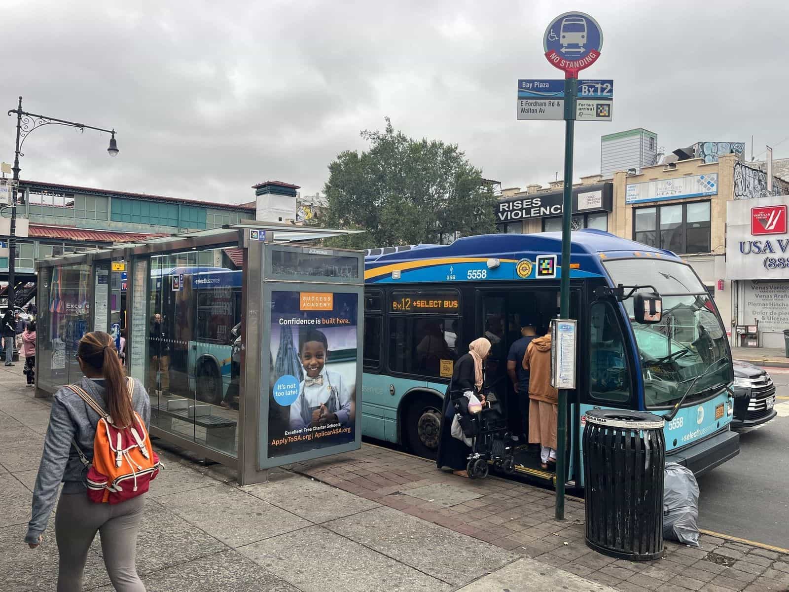 Pensilina della fermata Bx12-SBS «Bay Plaza · E Fordham Rd & Walton Av» con codice NaviLens accanto al cartello «For bus arrival»; in fondo, il bus 5558 con la porta aperta e i viaggiatori che salgono, compresa una persona in sedia a rotelle