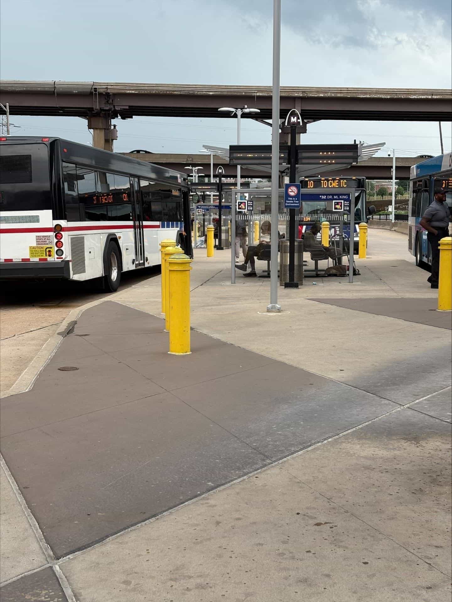 Blick auf das Civic Center Transit Center unter dem erhöhten MetroLink-Steg: zwei abgestellte MetroBusse — ein weißer mit rotem Streifen und der orangefarbenen LED-Anzeige „10 TO CWE TC" und ein blauer mit „32 DR. ML KING" —, in der Mitte ein Wartehäuschen mit schwarzem Dach, Metallbänken mit wartenden Fahrgästen, einer grauen Mülltonne, gelben Pollern und einem blauen „Bus Info"-Schild mit NaviLens-Code; braune Stahlsäulen und ein grauer Himmel im Hintergrund