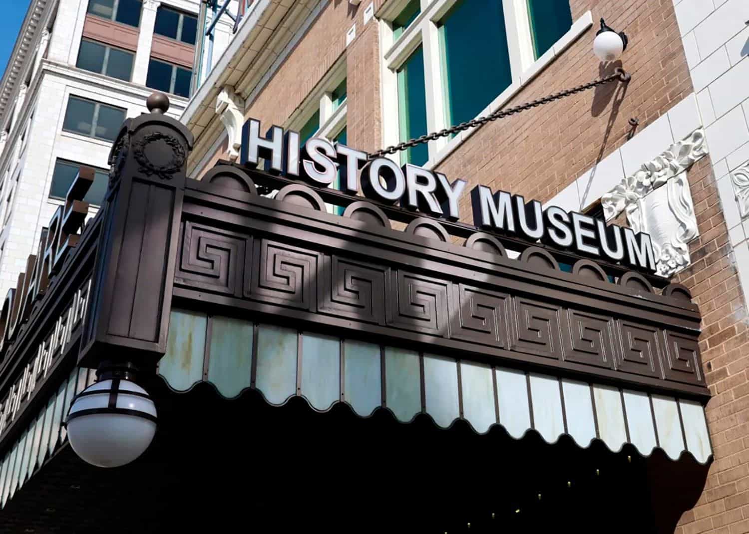 Exterior marquee of the History Museum on the Square in Springfield (Missouri): dark metal structure hanging from the brick facade of the Fox Theatre building with raised white “HISTORY MUSEUM” letters, scalloped edges, stained-glass panels and a classic spherical glass lamp