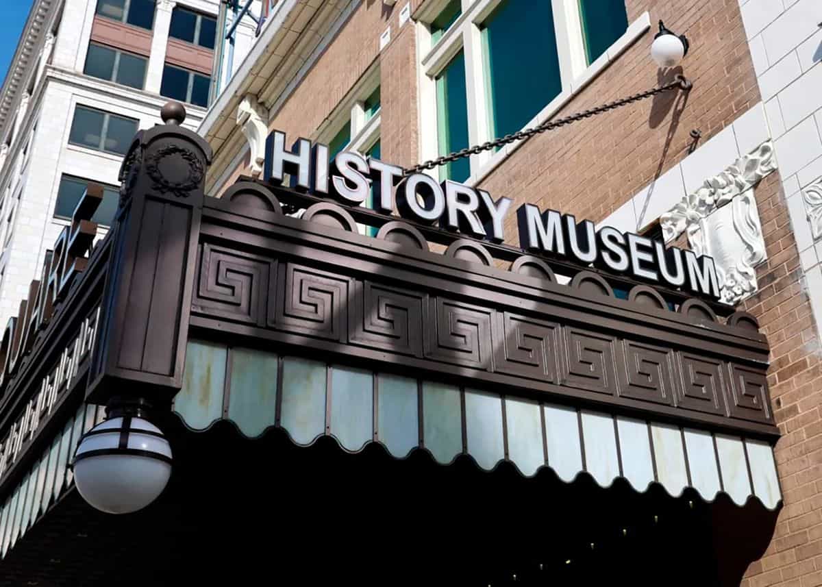Exterior marquee of the History Museum on the Square in Springfield (Missouri): dark metal structure hanging from the brick facade of the Fox Theatre building with raised white «HISTORY MUSEUM» letters, scalloped edges, stained-glass panels and a classic spherical glass lamp
