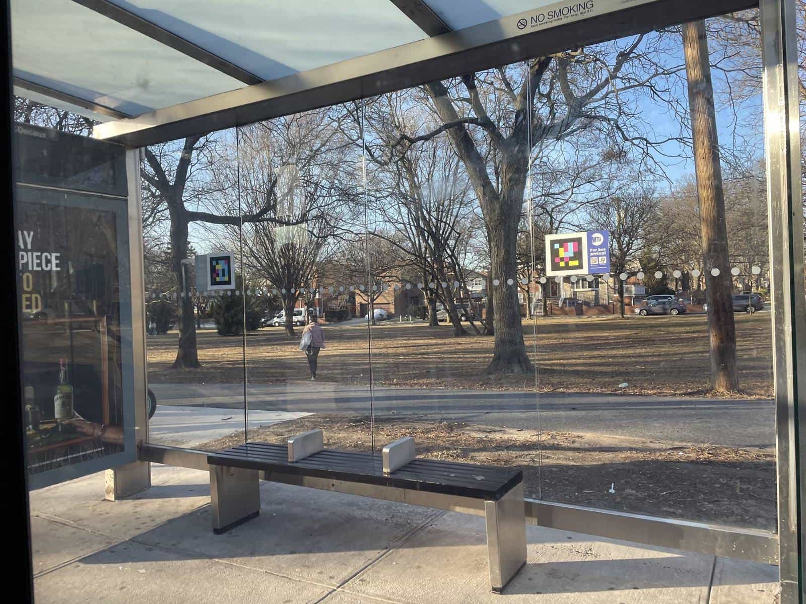 Inside a Bronx SBS shelter seen from the bench: glass panels, a park of bare trees behind and two NaviLens codes placed on the side panels