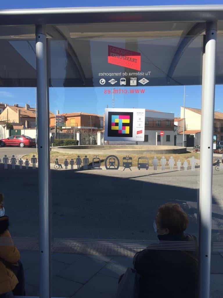 Cartel «Escanea con / Scan with» con un código NaviLens cuadrado a colores, iconos de las apps NaviLens y NaviLens GO, logo del CRTM y logo de Clear Channel pegado sobre el cristal de una marquesina