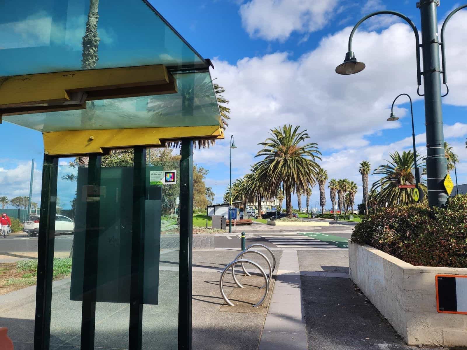 Pensilina di una fermata del tram con copertura in vetro verde-azzurro e travi gialle sul lungomare di Melbourne, con un cartello applicato all’interno del vetro —«Use NaviLens at this tram stop»— accanto a un codice NaviLens con cornice bianca; in fondo palme e la baia