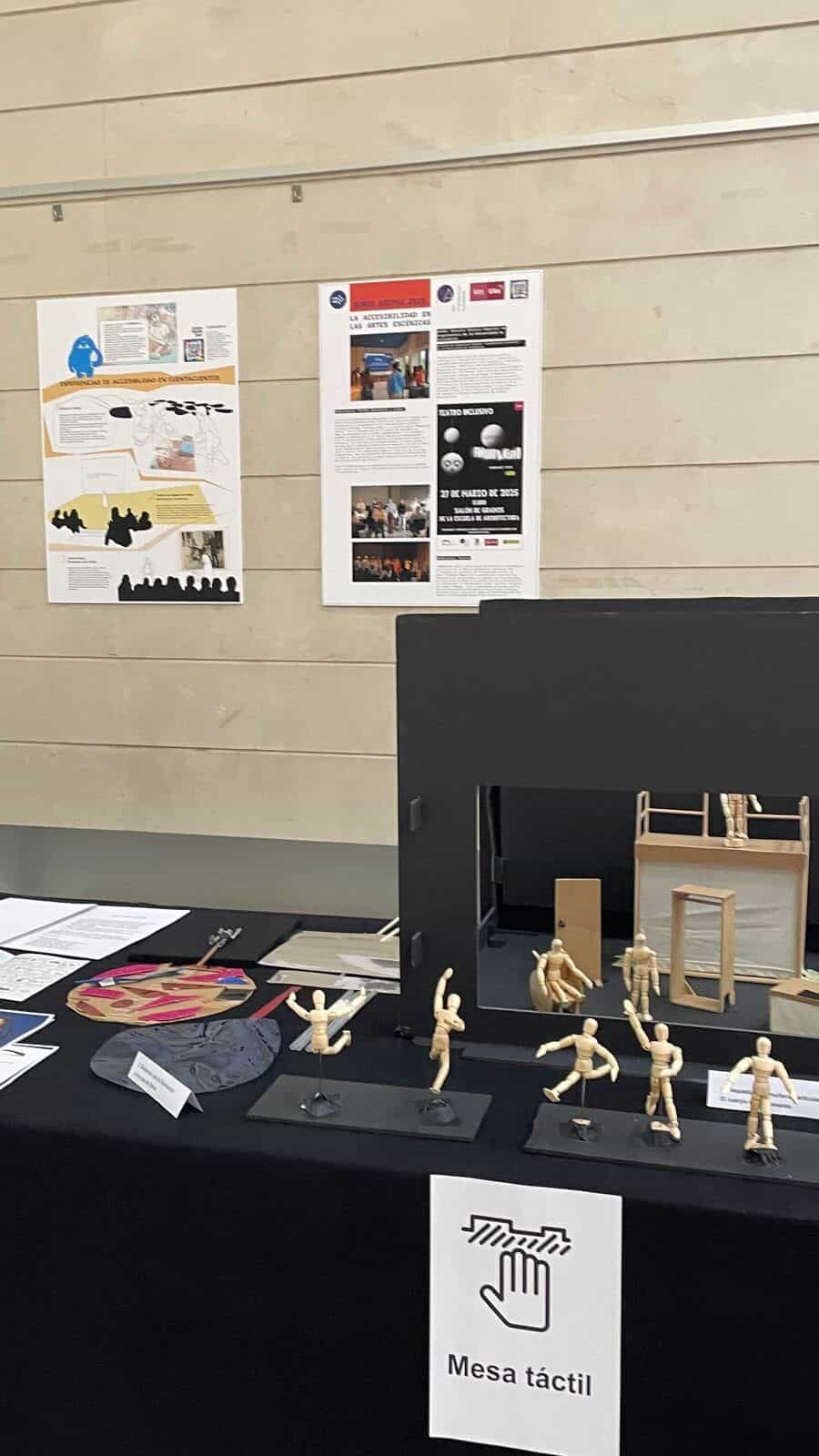 Tasttisch der Ausstellung „Somos ASEPAU 2025“ in der RESAD: Auf einer schwarzen Tischdecke inszenieren mehrere bewegliche Holzfiguren einen kleinen Chor und eine Theaterszene mit Miniatur-Bühnenvorhängen; im Vordergrund eine weiße Karte mit dem Piktogramm einer berührenden Hand und dem Text „Tasttisch“; im Hintergrund die Ausstellungstafeln und der NaviLens-Code an der Wand