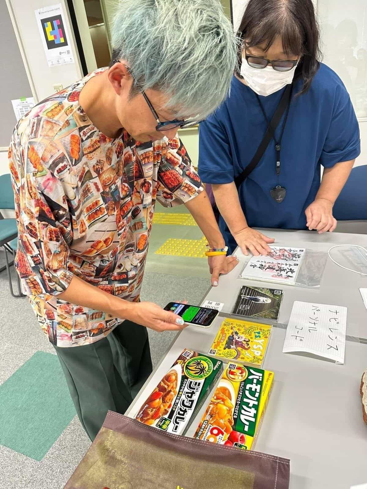 NaviLens カード、コードを貼った日本のカレー箱、手書きの「ナビレンス パーソナル コード」と書かれた札が並ぶテーブル