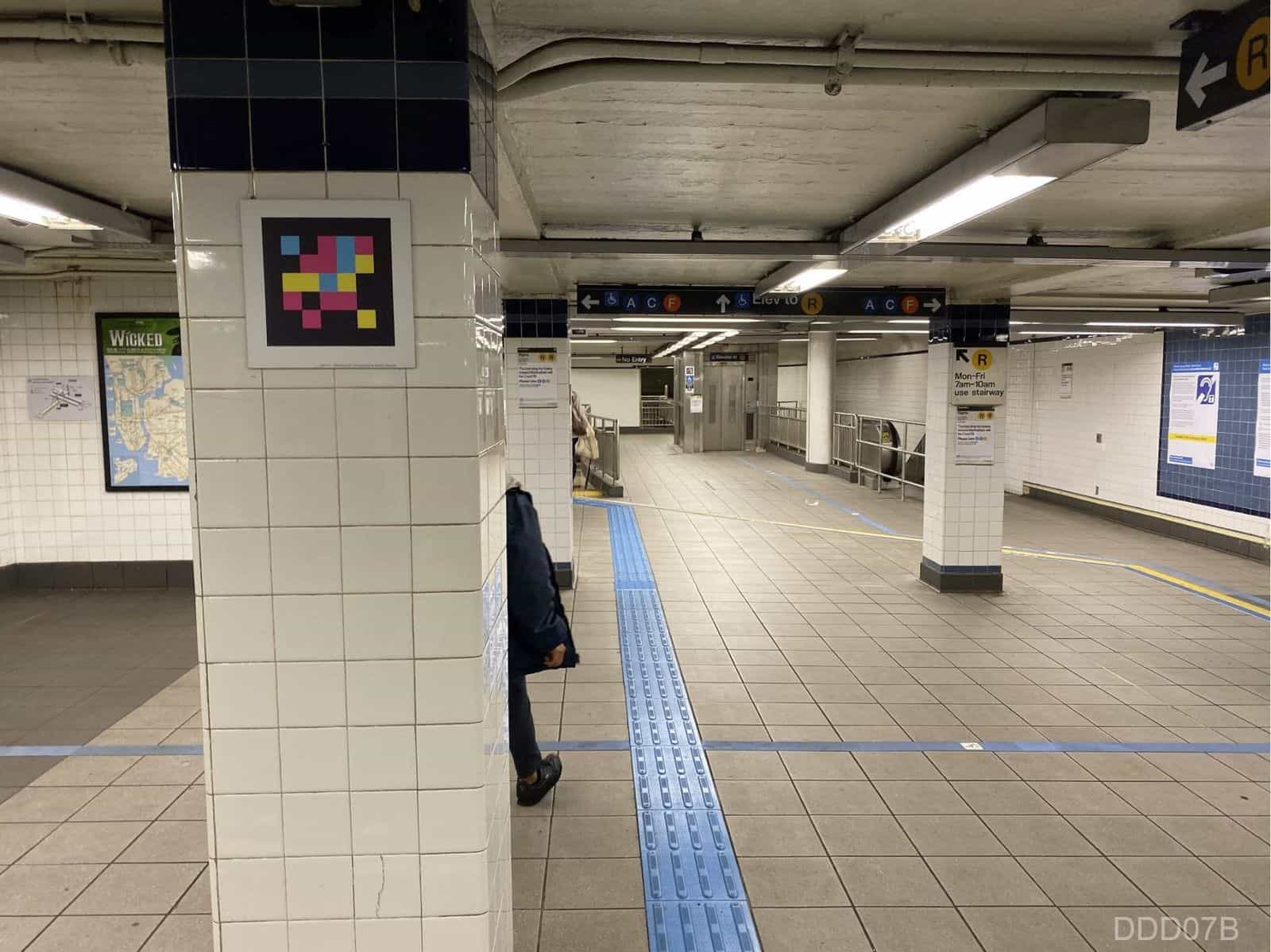 Jay St-MetroTech mezzanine: corridor with grey tiles and blue tactile guide-path. To the left, a white-tiled column with a NaviLens code and, in the background, hanging signs with arrows for A C F and «Elev to R»