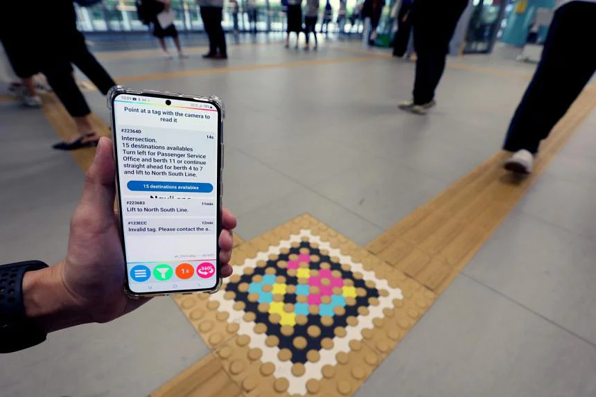 Hand holding a smartphone over a yellow tactile guide line at Woodlands ITH with a large NaviLens code on the floor; on screen the app shows “Intersection. 15 destinations available”