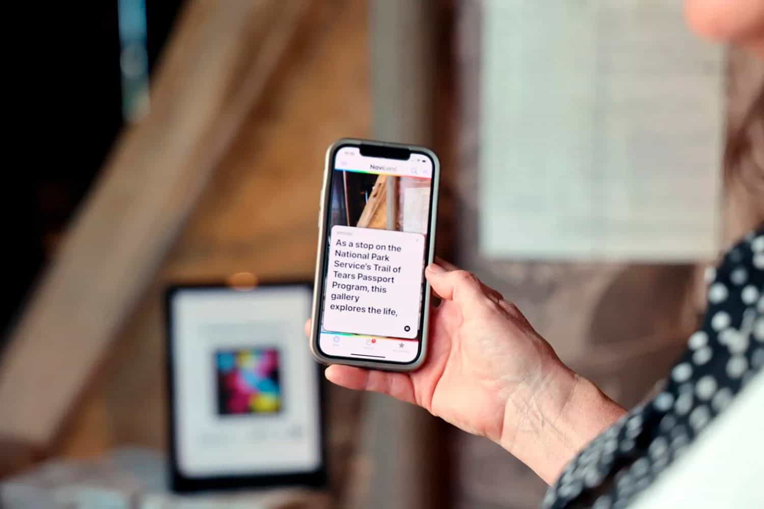 Hand of a visitor in a white polka-dot sleeve holding an iPhone in front of a museum panel: the NaviLens app on screen shows the Trail of Tears Passport Program label; in the blurred background, the wooden structure of a tipi