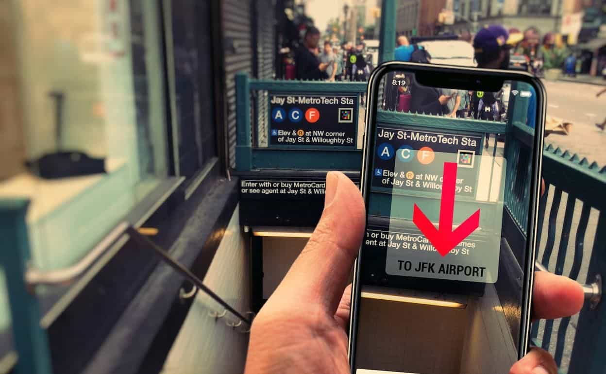 Subjektive Sicht auf der Straße vor dem Eingang der Station Jay St-MetroTech: eine Hand hält ein Handy, das über dem grünen Treppengeländer das schwebende AR-Schild «Jay St-MetroTech Sta · A C F · Elev & R at NW corner of Jay St & Willoughby St» mit einem dicken roten Pfeil nach unten und dem Label «TO JFK AIRPORT» zeigt