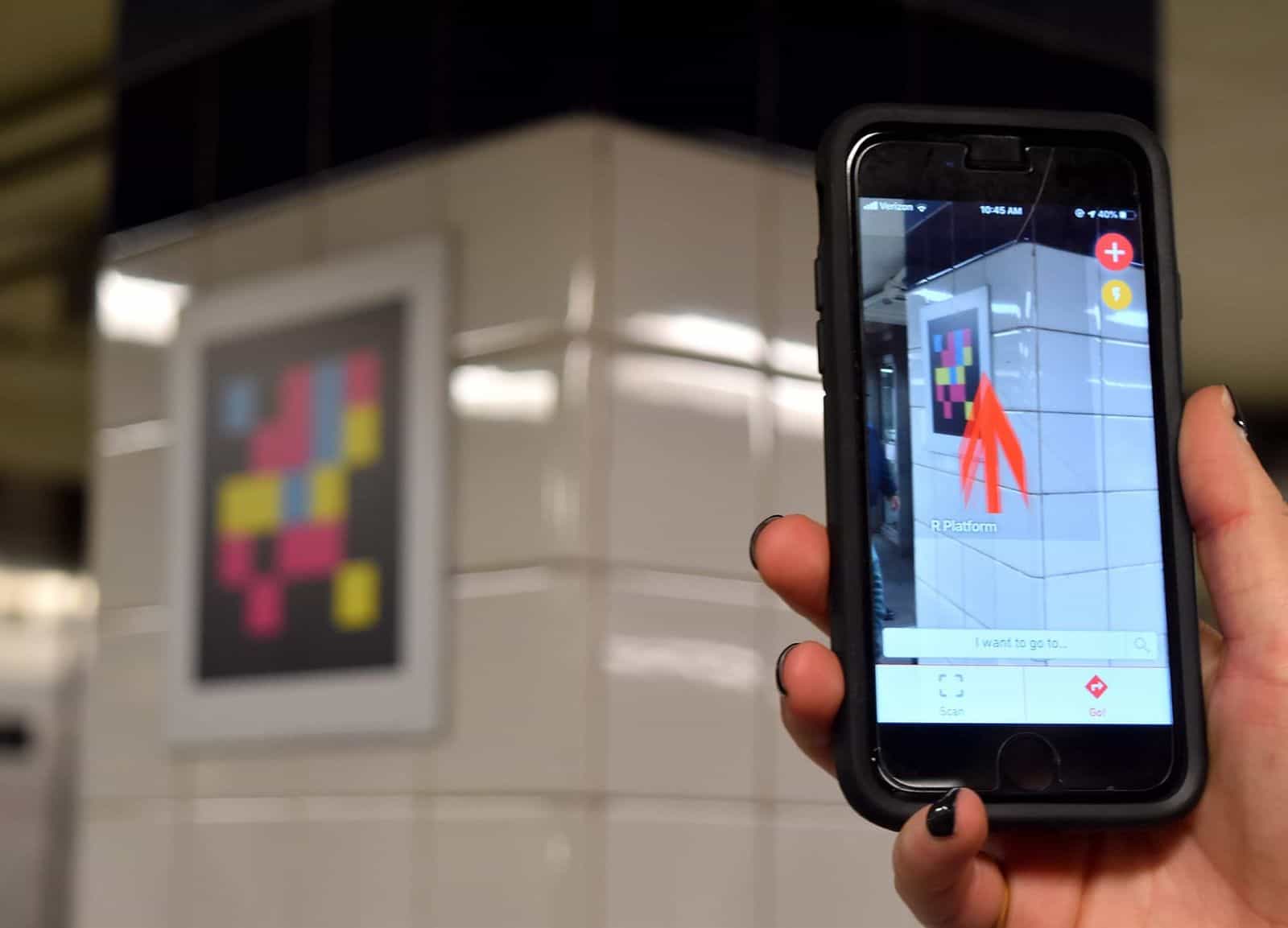 Hand mit iPhone und geöffneter NaviLens-App in einer U-Bahn-Station: die Kamera fokussiert einen NaviLens-Code an einer weiß gefliesten Säule, und über dem realen Bild zeigt ein dicker oranger Pfeil mit dem Text «R Platform» den Weg zum Bahnsteig der Linie R
