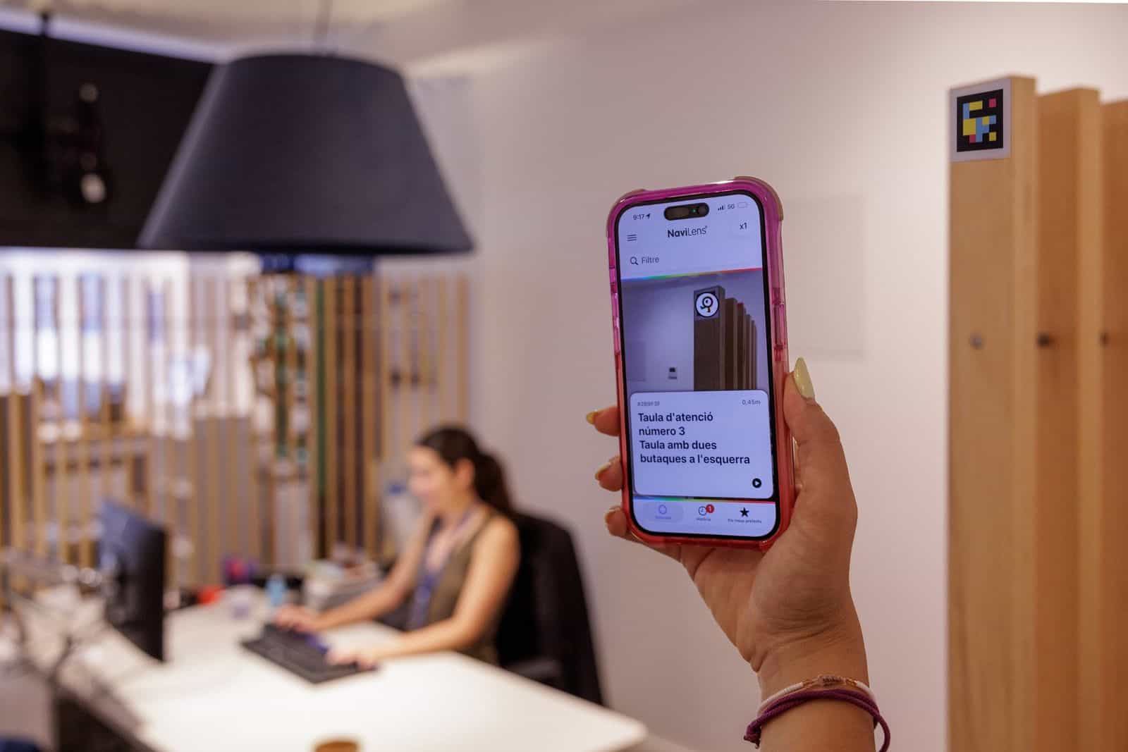 NaviLensアプリでAigües de Barcelonaのカウンター情報をカタルーニャ語で読み上げているスマートフォンを手に持つ様子。