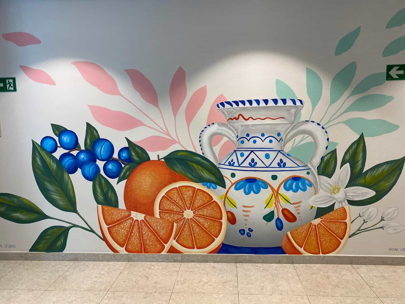 Handgemaltes Wandbild am ibis Styles Sevilla: große weiße Keramikkanne mit blauen andalusischen Motiven, umgeben von Orangen, Orangenspalten und dekorativem Blattwerk