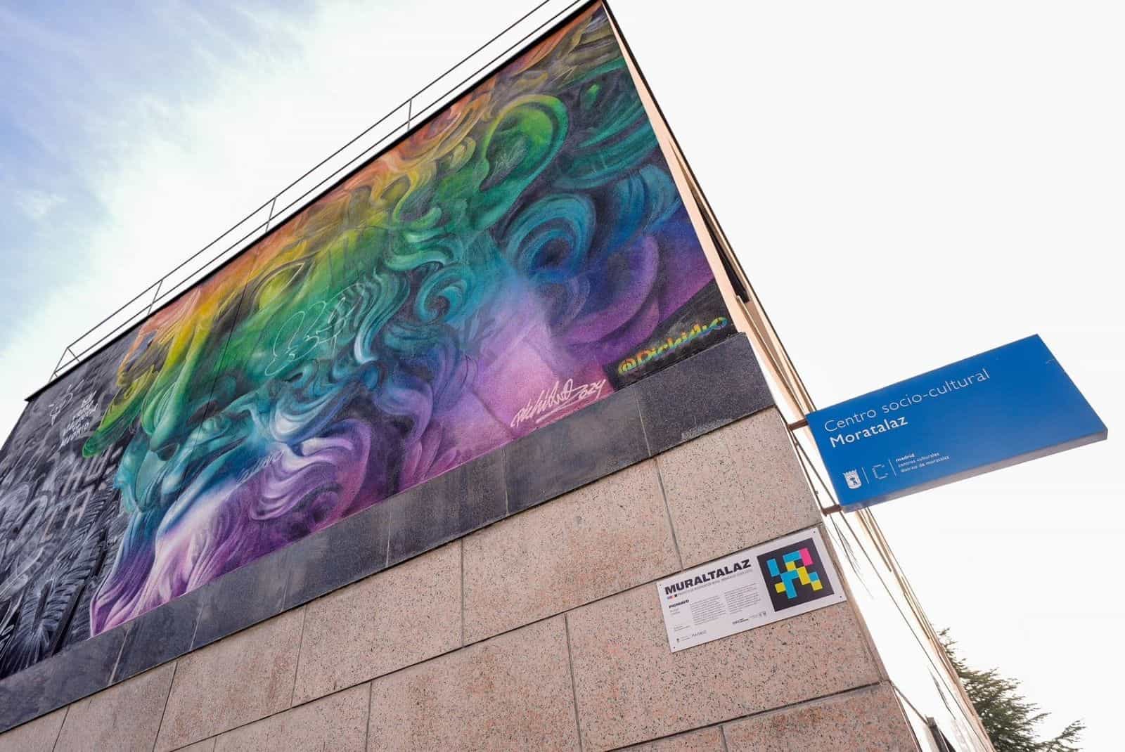 Murale di Pichiavo sulla facciata del centro socio-culturale Moratalaz, con targa bianca «MURALTALAZ» e codice NaviLens a colori accanto al granito.