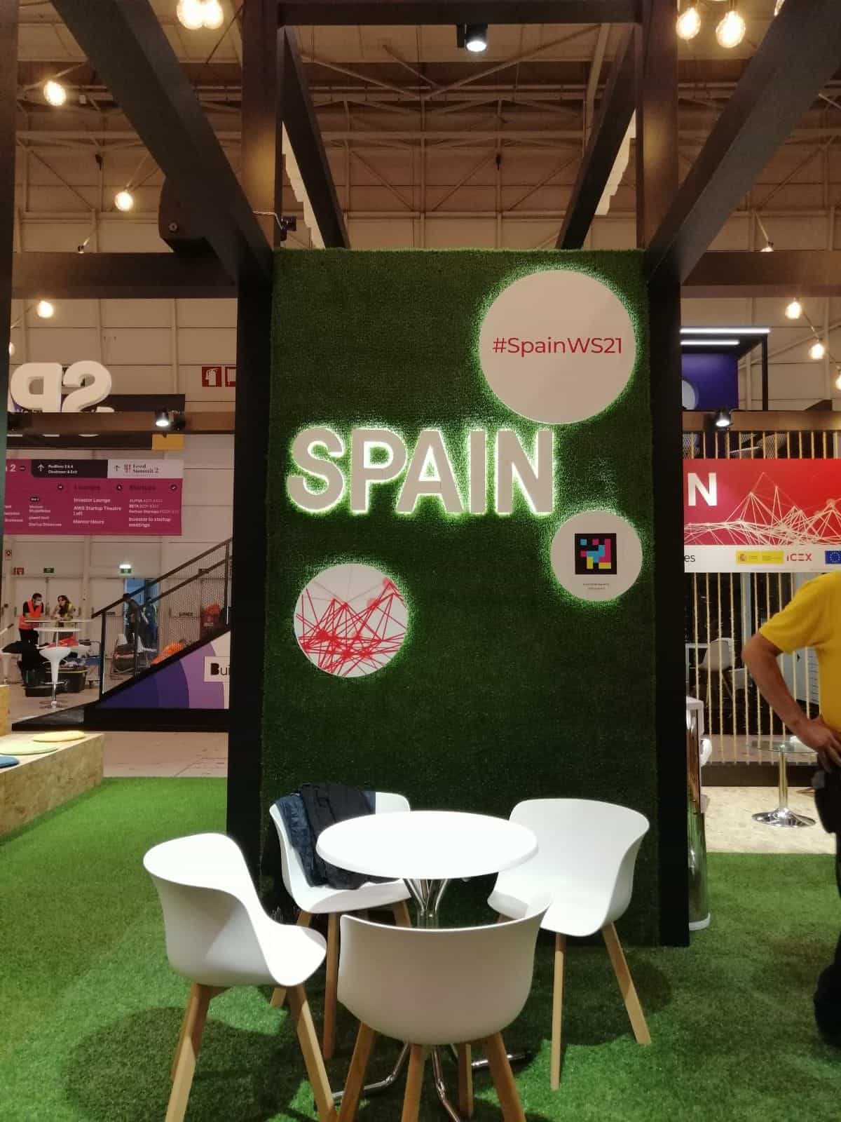 Muro de césped artificial del Pabellón de España en Web Summit 2021: en el centro, el rótulo luminoso «SPAIN» en blanco; en círculos blancos a la derecha, el hashtag «#SpainWS21» y un código NaviLens cuadrado a colores; a la izquierda, una ilustración roja de la red de contactos. Delante, mesa redonda blanca y sillas estilo Eames sobre césped verde