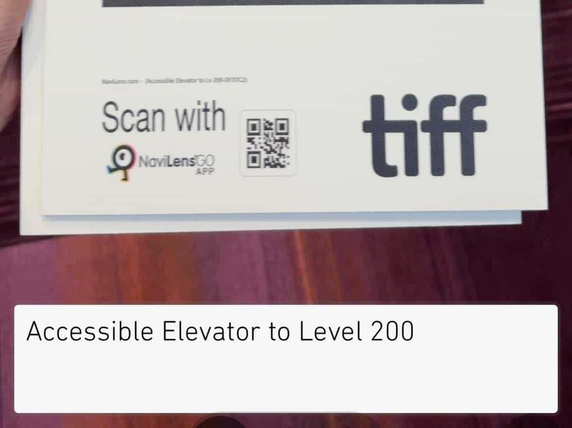 The NaviLens GO app reading a printed poster held in one hand: below the code are the line “Scan with NaviLens GO” and the TIFF logo, and at the bottom the app shows the text “Accessible Elevator to Level 200.”