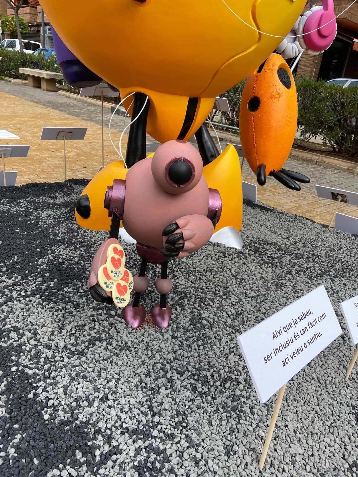 Dettaglio di un ninot dei bambini della falla «Inclusivament»: una figura tiene un mazzetto di spille gialle con cuori rossi e la parola «INCLUSIVAMENT», emblema del progetto della commissione Cronista Vicent Beguer i Esteve di Torrent