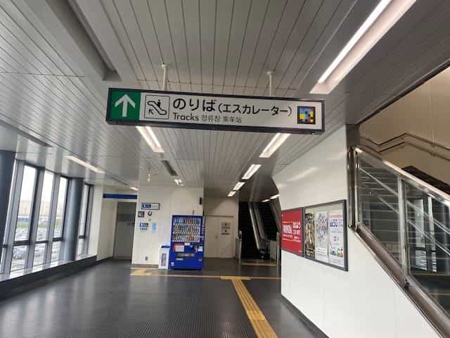 神戸空港駅の緑色吊り下げサイン「のりば(エスカレーター)/ Tracks」、上向き矢印、エスカレーター絵文字、NaviLensタグ。奥には上り方向のエスカレーターと飲料自販機