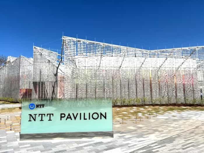 NTTパビリオン「PARALLEL TRAVEL」の半透明ファサード、青空を背景にした銀色のチューブパネルと前景の企業ロゴ