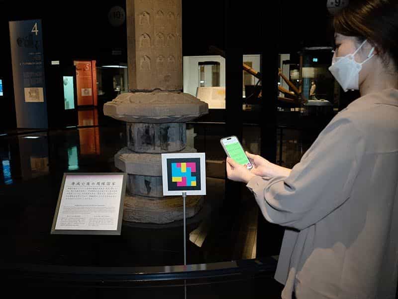 文化交流展示室で、石造の塔とキャプションの横に設置されたNaviLensコードを、来館者がスマートフォンでスキャンしている様子