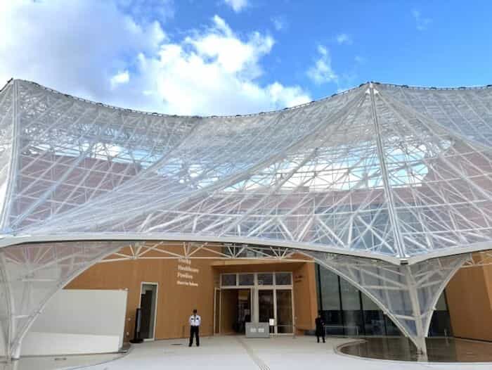 白い張力膜屋根を持つ大阪ヘルスケアパビリオン「Nest for Reborn」、構造メッシュの下に木製の正面入口